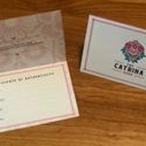 Pode incluir: Um cart&atilde;o dobrado com um padr&atilde;o floral rosa e branco no interior e um cart&atilde;o azul com um logotipo de rosa e o texto "The Catrina" no exterior.