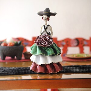 Pode incluir: Uma figura de mulher esqueleto vestida com um vestido verde, branco e vermelho e um chap&eacute;u preto. Ela est&aacute; segurando um buqu&ecirc; de rosas vermelhas.