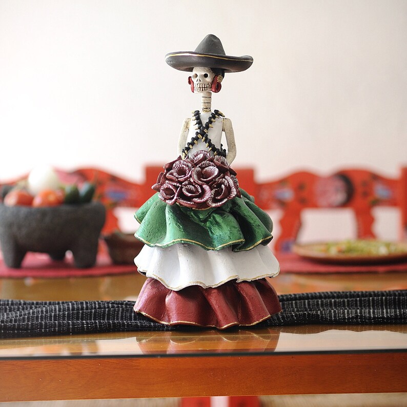 Pode incluir: Uma figura de mulher esqueleto vestida com um vestido verde, branco e vermelho e um chap&eacute;u preto. Ela est&aacute; segurando um buqu&ecirc; de rosas vermelhas.