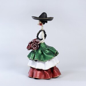 Pode incluir: Uma figura de mulher esqueleto do Dia dos Mortos com um vestido verde, branco e vermelho e um chap&eacute;u preto. Ela est&aacute; segurando um buqu&ecirc; de flores vermelhas.