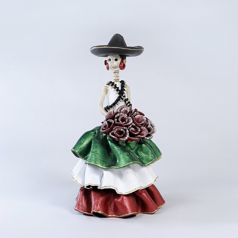 Pode incluir: Uma figura de mulher esqueleto do Dia dos Mortos usando um sombrero preto, um vestido verde, branco e vermelho, e segurando um buqu&ecirc; de flores vermelhas.