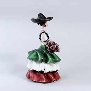 Pode incluir: Uma figura de mulher esqueleto do Dia dos Mortos com um chap&eacute;u preto, um vestido verde, branco e vermelho, e segurando um buqu&ecirc; de flores vermelhas.