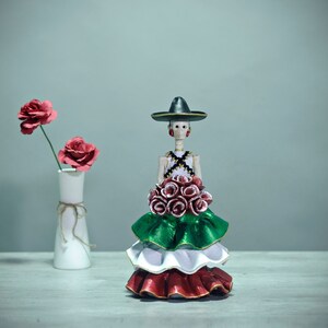 Pode incluir: Uma figura do Dia dos Mortos vestindo um sombrero preto e um vestido verde, branco e vermelho segurando um buqu&ecirc; de flores vermelhas.