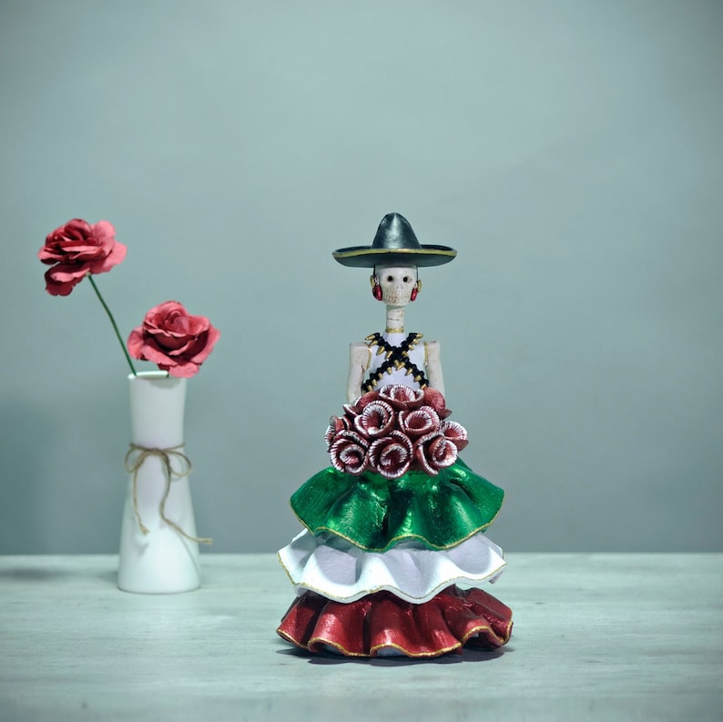 Pode incluir: Uma figura do Dia dos Mortos vestindo um sombrero preto e um vestido verde, branco e vermelho segurando um buqu&ecirc; de flores vermelhas.