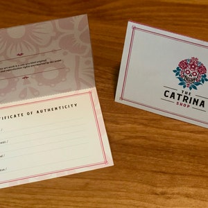 Op de afbeelding: Een echtheidscertificaat met een roze en wit bloemenontwerp. Het certificaat is voor een kunstwerk. De tekst "Echtheidscertificaat" staat in het zwart bovenaan het certificaat. De tekst "Titel van het kunstwerk", "Naam van de kunstenaar", "Medium / Materialen", "Afmetingen", "Jaar van voltooiing" en "Handtekening van de kunstenaar" staat in het zwart onder de titel. Het certificaat is doormidden gevouwen. De achterkant van het certificaat heeft een roze en wit bloemenontwerp. De tekst "Dit kunstwerk is een uniek origineel. Alle auteursrechten en reproductierechten zijn voorbehouden aan de kunstenaar." staat in het zwart onderaan het certificaat.