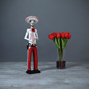 Op de afbeelding: Een wit skeletfiguur dat een rood-witte sombrero en een rood-wit gepatroneerd pak draagt. Het figuur houdt zijn handen voor zijn borst. Op de achtergrond staat een vaas met rode tulpen.