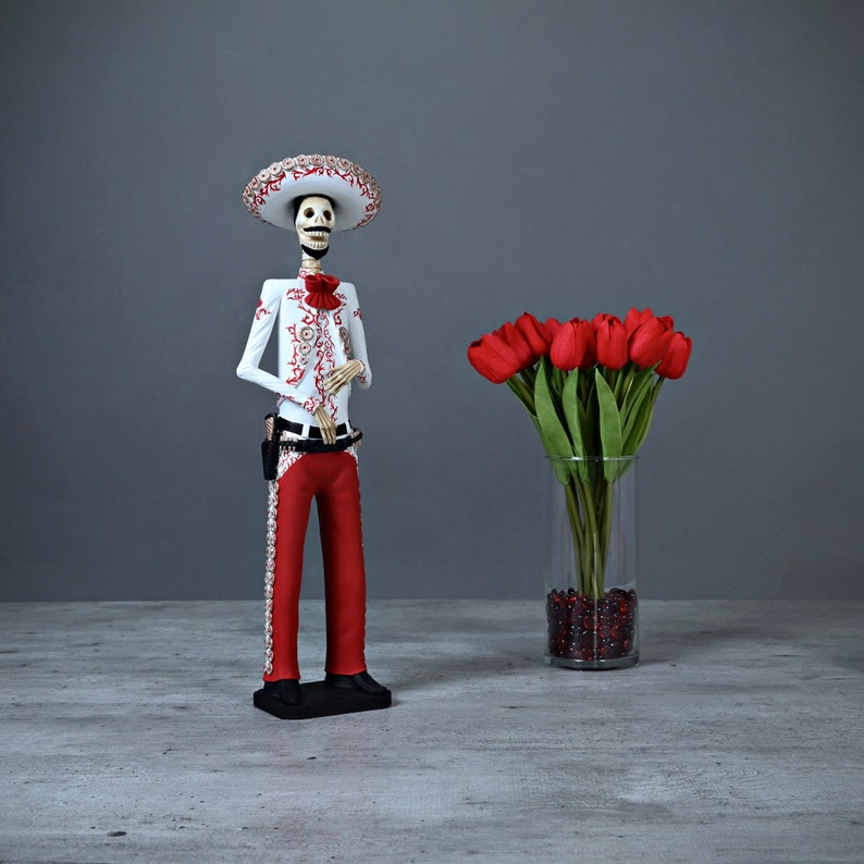 Op de afbeelding: Een wit skeletfiguur dat een rood-witte sombrero en een rood-wit gepatroneerd pak draagt. Het figuur houdt zijn handen voor zijn borst. Op de achtergrond staat een vaas met rode tulpen.