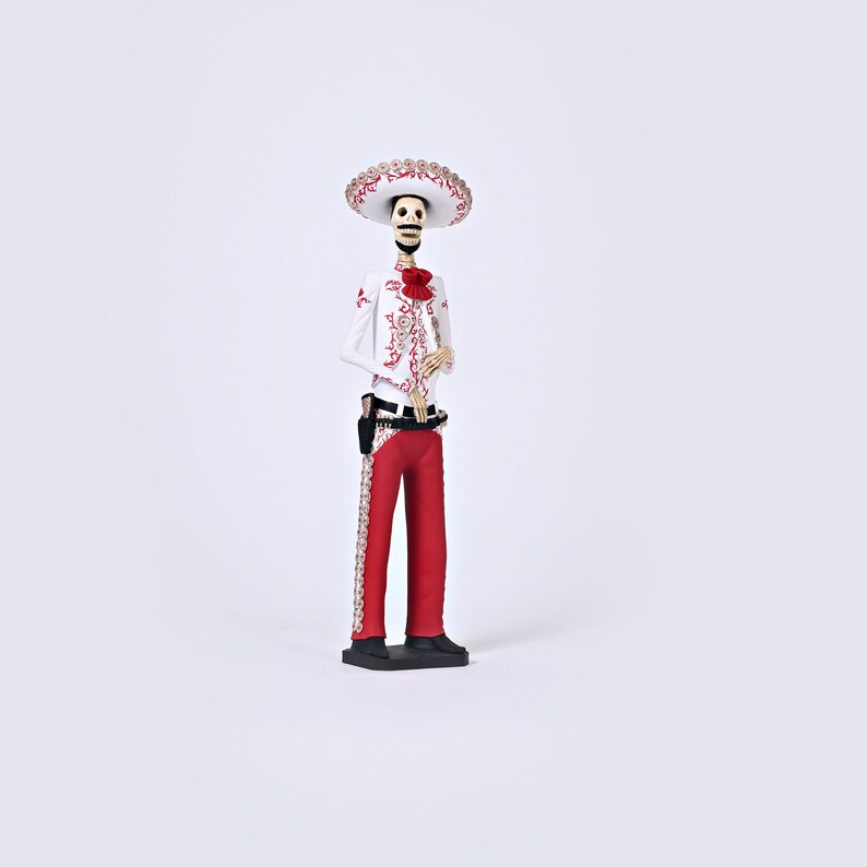 Op de afbeelding: Een wit skeletfiguur dat een rood-wit geborduurd pak draagt, een zwarte riem en een brede sombrero met rode en gouden versieringen. Het figuur staat op een zwarte basis.