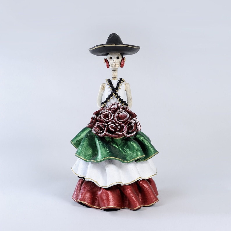 Pode incluir: Uma figura de esqueleto do Dia dos Mortos vestindo um sombrero preto, uma blusa branca e uma saia de camadas verde, branca e vermelha com rosas vermelhas.