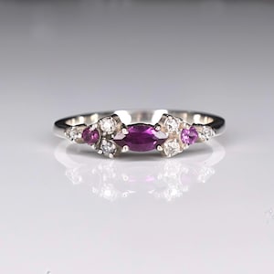 May include: A silver ring featuring a marquise-cut, deep purple gemstone at the center, flanked by clusters of smaller, round white and pink gemstones. The ring's band is thin and polished, reflecting light.
