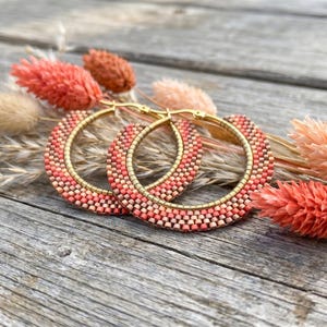 May include: A pair of hoop earrings featuring a beaded design in shades of coral, peach, and cream. The beads are arranged in a gradient pattern along the outer edge of the gold-toned metal hoops, creating a textured appearance on the circular frames.
