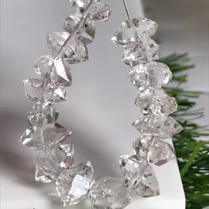 Peut inclure: Un collier de cristaux de quartz bruts, transparents et à double terminaison, enfilés sur un fil métallique argenté. Chaque cristal présente des formes géométriques naturelles à facettes avec des inclusions internes variées.