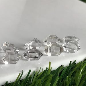 Peut inclure: Un ensemble de huit petits cristaux de quartz transparents à double terminaison, présentant des facettes hexagonales naturelles. Ces pierres incolores sont disposées en groupe, mettant en valeur leurs pointes et leur structure prismatique.