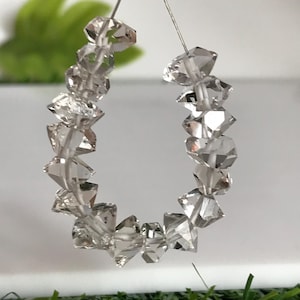 Peut inclure: Un collier de perles transparentes, facettées et incolores disposées en forme de U. Chaque perle présente de multiples surfaces géométriques qui reflètent la lumière, créant un aspect scintillant. Les perles sont enfilées sur un fil métallique.