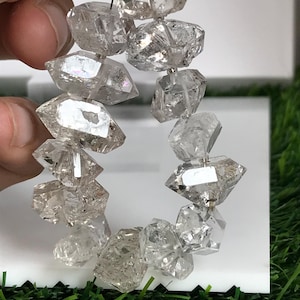 Peut inclure: Un collier de cristaux de quartz bruts et transparents aux formes irrégulières et facettées, présentant des inclusions internes. Les pierres sont enfilées sur un fil fin, offrant un aspect translucide semblable à du verre avec des variations naturelles.