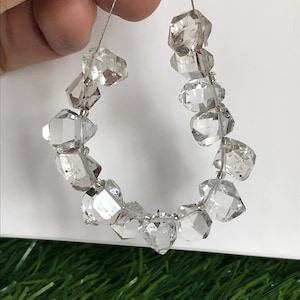 Peut inclure: Un collier de cristaux de quartz transparents, bruts et facettés, enfilés sur un fil fin de couleur argentée. Les pierres sont de forme irrégulière, présentant une transparence naturelle et des surfaces réfléchissantes sur l'ensemble.