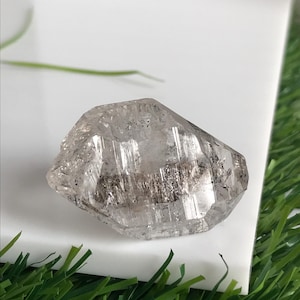 Peut inclure: Un cristal de quartz transparent à facettes multiples présentant des inclusions internes visibles et des dépôts minéraux sombres en forme d'aiguilles, concentrés en une bande horizontale au centre de la pierre, avec une structure naturelle.