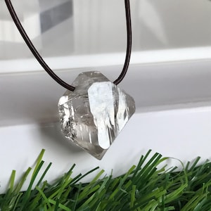 Op de afbeelding: Een heldere, veelzijdige kwartskristallen hanger hangend aan een dun, donkerbruin leren koord. Het kristal heeft een geometrische, diamantachtige vorm met zichtbare interne breuklijnen en natuurlijke facetten die het licht weerkaatsen.