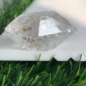 Könnte beinhalten: Ein klarer, facettierter Quarzkristall mit doppelt endender Form. Der Stein weist interne Risse und kleine, hellbraune mineralische Einschlüsse in seiner gesamten Struktur auf, die das Licht auf seinen polierten, geometrischen Oberflächen reflektieren.