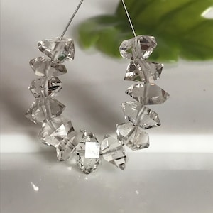 Peut inclure: Un collier de onze cristaux de quartz transparents, facettés et à double terminaison, disposés en demi-cercle. Chaque pierre présente une structure géométrique pointue avec de multiples surfaces réfléchissantes et des inclusions visibles.
