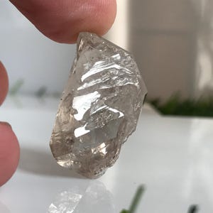 Peut inclure: Un cristal de quartz fumé brut et translucide avec une surface dentelée et à facettes multiples. La pierre présente une teinte brun pâle grisâtre avec des fractures internes et des bords tranchants qui captent la lumière, créant un aspect cristallin.