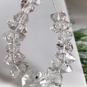 Peut inclure: Un collier de cristaux de quartz transparents, facettés et à double terminaison, disposés en forme de U. Chaque pierre présente une géométrie hexagonale pointue avec des inclusions naturelles et une grande transparence, enfilée sur un fil argenté.