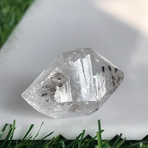 Peut inclure: Un cristal de quartz transparent à double terminaison présentant des facettes naturelles et des inclusions internes. La pierre a une forme géométrique hexagonale avec des stries visibles sur sa surface, reflétant la lumière à travers sa structure transparente.
