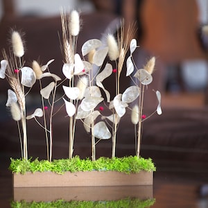 Peut inclure: Composition florale décorative avec du blé séché, des queues de lapin blanches et duveteuses et des plantes délicates en dollars d'argent. L'arrangement est placé dans une boîte en bois rectangulaire remplie de mousse verte vibrante, ornée de petites baies rouges.