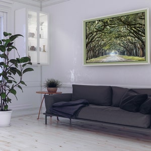May include: A living room interior with a gray sofa, a white potted plant, a small side table, and a framed photograph of a tree-lined path. The photograph is hanging on a white wall.