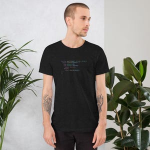 May include: Dark gray t-shirt with colorful code text. The text is in a teal, blue, and orange font. The shirt is short-sleeved and has a crew neck. The model is wearing the shirt.