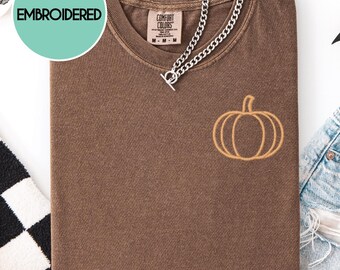 May include: A brown t-shirt with an embroidered pumpkin design. The shirt has a V-neck and is accessorized with a silver chain necklace. The words "Embroidered" and "Custom Thread Color" are displayed on the image.
