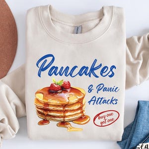 May include: A beige Gildan Sand sweatshirt with the words "Pancakes & Panic Attacks" in blue script. The graphic shows a stack of pancakes with syrup and strawberries. A brown hat and blue jeans are also in the image.