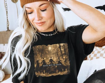 May include: Black Bella Canvas t-shirt featuring a graphic of four witches in a forest with the text "Girls Will Be Girls." The shirt is worn with a brown beanie and black pants. The image is taken indoors.