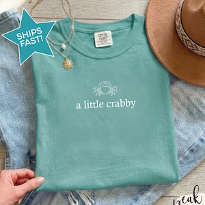 Op de afbeelding: Een zeeschuimgroen T-shirt met korte mouwen, voorzien van een witte lijntekening van een krab en de tekst a little crabby. Het shirt ligt op een lichtblauwe spijkerbroek en wordt vergezeld door een beige vilten hoed en een goudkleurige ketting.