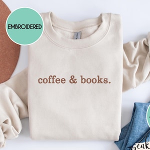 May include: A beige embroidered sweatshirt with the text "coffee & books." A brown hat and blue jeans are next to the sweatshirt. The sweatshirt is displayed on Gildan Sand. The thread color can be customized.