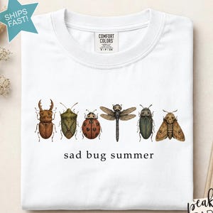 May include: A white crew neck t-shirt featuring a horizontal row of six detailed insect illustrations including a beetle, a stink bug, a ladybug, a dragonfly, a dark beetle, and a moth, with the text "sad bug summer" printed in black below.