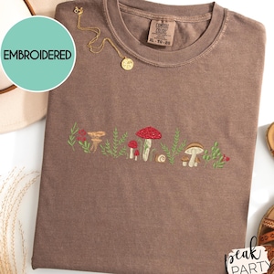 May include: Brown t-shirt with an embroidered mushroom design. The design features red, brown, and white mushrooms with green foliage. A gold necklace and a beige hat are also visible.
