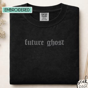 May include: A black crewneck t-shirt featuring the embroidered text future ghost in a gothic font centered on the chest. The shirt includes a Comfort Colors brand label on the inside collar and the word EMBROIDERED in a light blue box.