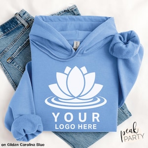 May include: A light blue hoodie with a white lotus flower design and the text "YOUR LOGO HERE". The hoodie is displayed on a pair of blue jeans. The text "Displayed on Gildan Carolina Blue" is at the bottom left.