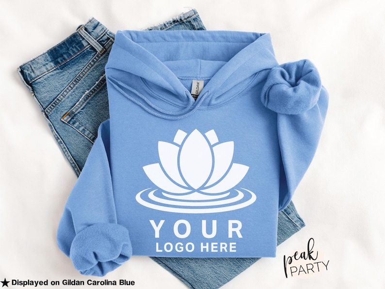 May include: A light blue hoodie with a white lotus flower design and the text "YOUR LOGO HERE". The hoodie is displayed on a pair of blue jeans. The text "Displayed on Gildan Carolina Blue" is at the bottom left.
