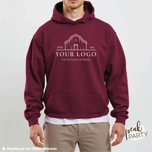 May include: Maroon hoodie with a front pocket and a drawstring hood. The front of the hoodie features a white logo with the text "YOUR LOGO" and "YOUR SLOGAN HERE" below a barn illustration. The text "ESTD 2022" is also included.
