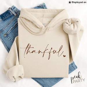 May include: A cream-colored hooded sweatshirt with the word "thankful" in a brown script font, accompanied by a small heart. The sweatshirt is displayed on top of blue denim jeans. The text "Displayed on Gildan Sand" is in the top right corner.