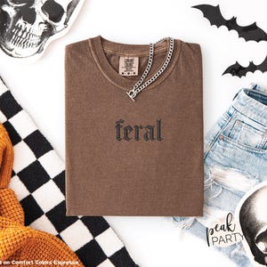 May include: A brown t-shirt with the word "feral" embroidered on the front, paired with a silver chain necklace. The image includes denim shorts, decorative bats, and skull-themed accessories, suggesting a Halloween or alternative aesthetic.