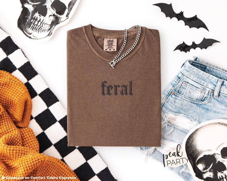 May include: A brown t-shirt with the word "feral" embroidered on the front, paired with a silver chain necklace. The image includes denim shorts, decorative bats, and skull-themed accessories, suggesting a Halloween or alternative aesthetic.