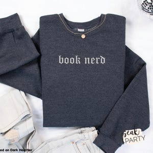Op de afbeelding: Donkergrijs sweatshirt met de geborduurde tekst "book nerd" in het wit. Een gouden ketting met een hanger ligt op het sweatshirt. Lichte jeans zijn ook zichtbaar. Het sweatshirt is opgevouwen.