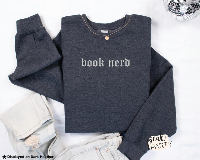 Op de afbeelding: Donkergrijs sweatshirt met de geborduurde tekst "book nerd" in het wit. Een gouden ketting met een hanger ligt op het sweatshirt. Lichte jeans zijn ook zichtbaar. Het sweatshirt is opgevouwen.