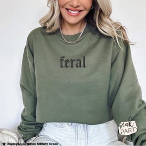 May include: Olive green sweatshirt with the word "feral" embroidered in black gothic lettering. The sweatshirt is displayed on a person wearing a silver chain necklace and light-wash jeans.