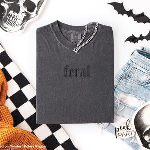 May include: A dark gray t-shirt with the word "feral" embroidered on the front. A silver chain necklace is draped over the shirt. The image includes denim shorts, skull decorations, and black bat cutouts, suggesting a Halloween theme.
