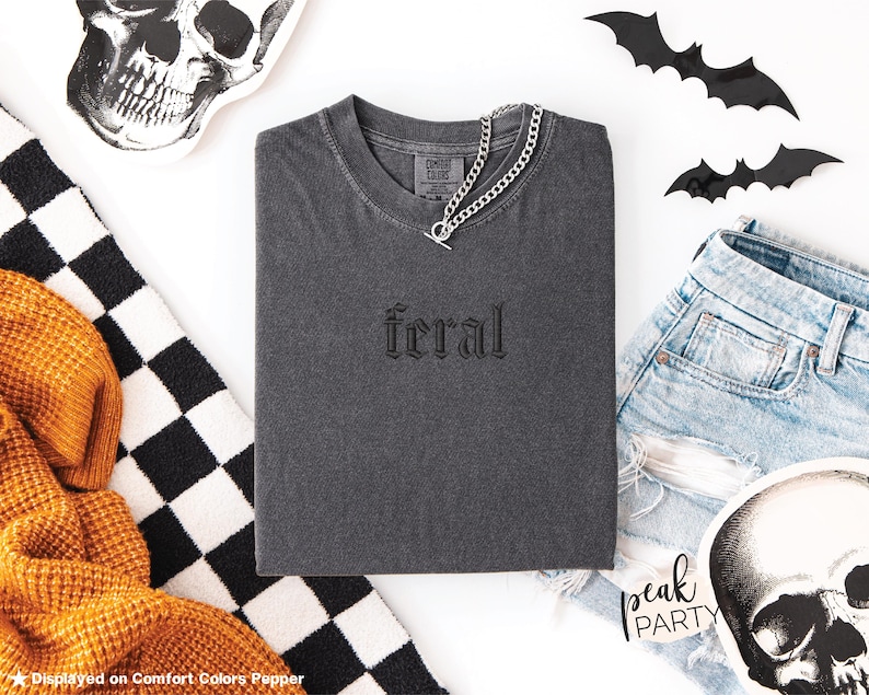 May include: A dark gray t-shirt with the word "feral" embroidered on the front. A silver chain necklace is draped over the shirt. The image includes denim shorts, skull decorations, and black bat cutouts, suggesting a Halloween theme.