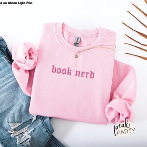 Op de afbeelding: Een lichtroze sweatshirt met de geborduurde tekst "book nerd" in een gotisch lettertype. Het sweatshirt wordt getoond met een spijkerbroek met distress details en een delicate ketting. De woorden "peak party" staan rechtsonder.